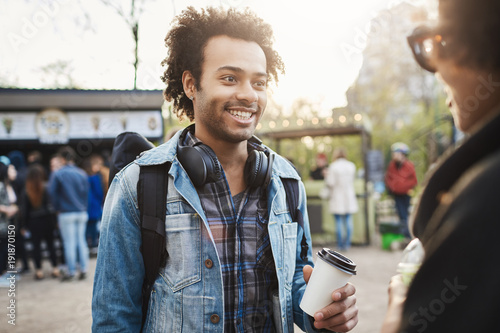 Outdoor portrait of stylish upbeat african-american male with bristle and afro hairstyle holding cup of coffee and smiling broadly while talking to attractive woman in park  asking for her number.