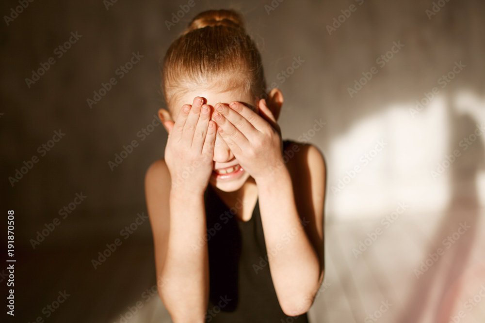 Close-up portrait of a little adorable young ballerina in black sportwear smiles and closes her eyes with her hands because of the bright sunlight Close-up portrait of a little adorable young ballerina in black sportwear smiles and closes her eyes with her hands because of the bright sunlight