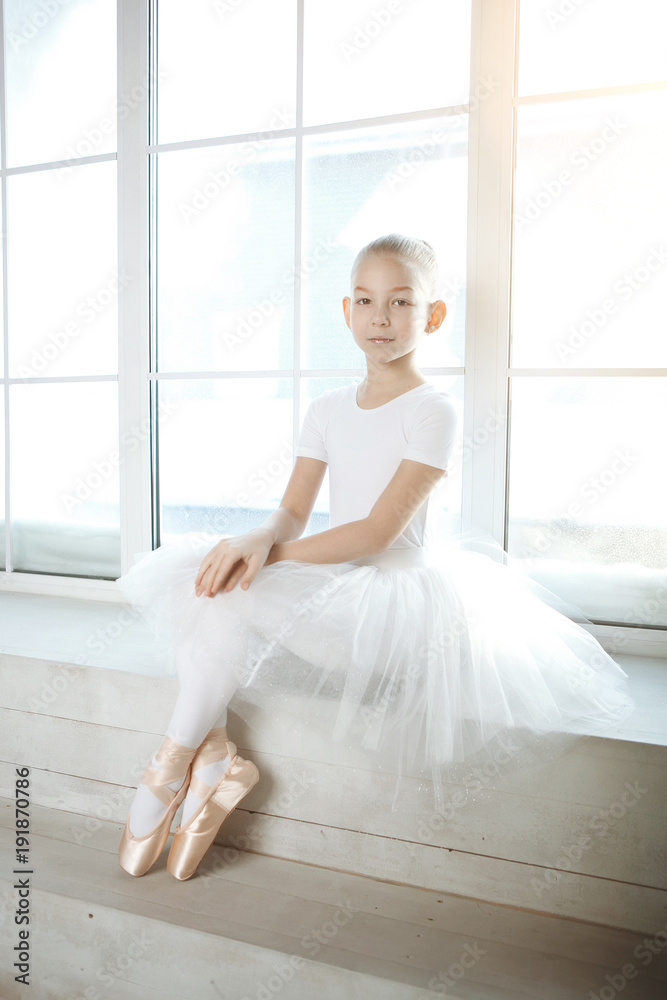 A little adorable young ballerina in white dress and point shoes sitting on the window background