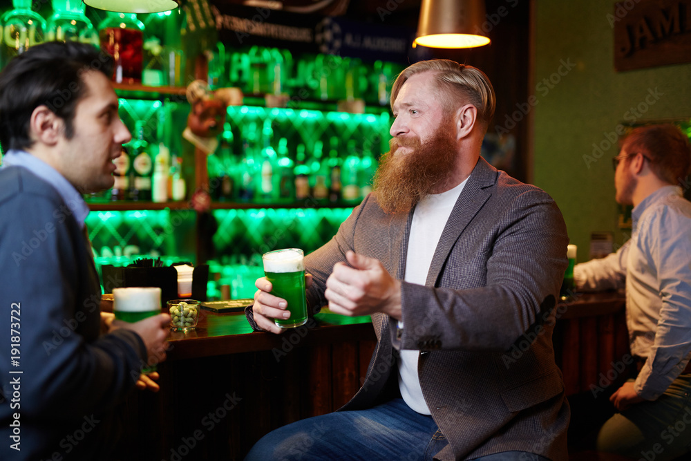 Bearded man and his friend with glasses of beer relaxing in pub and having talk