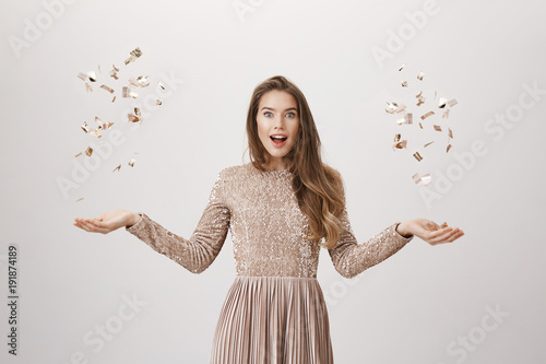 Surprised attractive young caucasian woman in evening dress tossing up confetti with both hands while glancing with opened mouth at camera over gray background. Girl on celebration photo