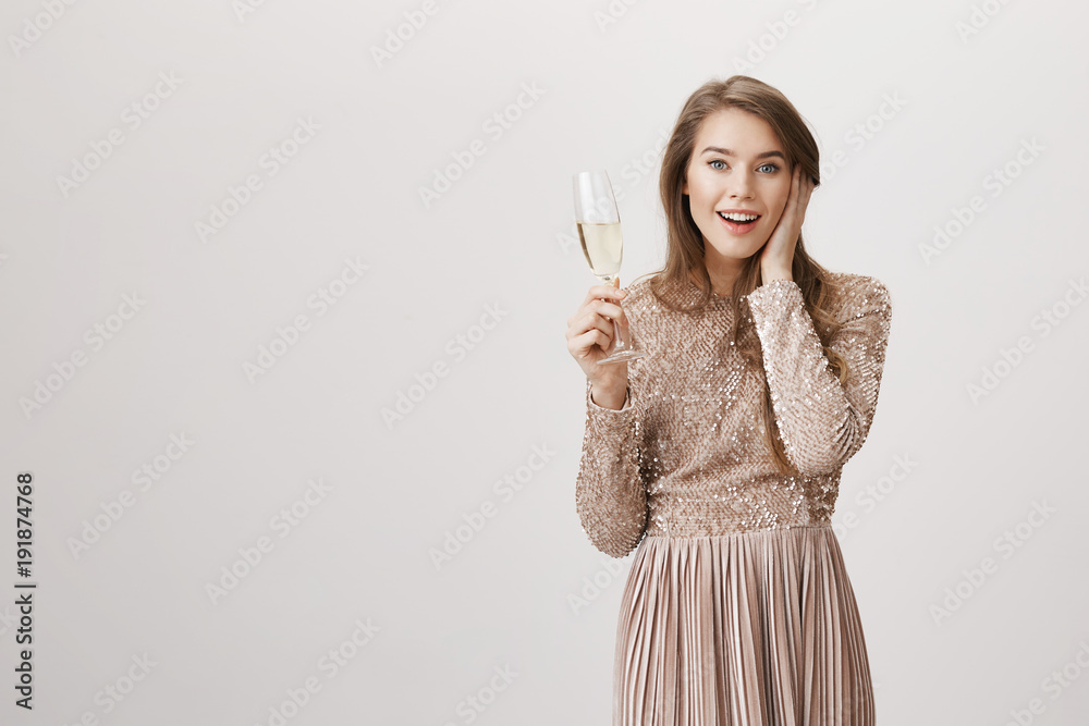 Surprised cute caucasian woman holding hand on cheek while standing in evening dress with glass of champagne, glancing at camera over gray background. Girl is shocked boyfriend proposed on party Surprised cute caucasian woman holding hand on cheek while standing in evening dress with glass of champagne, glancing at camera over gray background. Girl is shocked boyfriend proposed on party