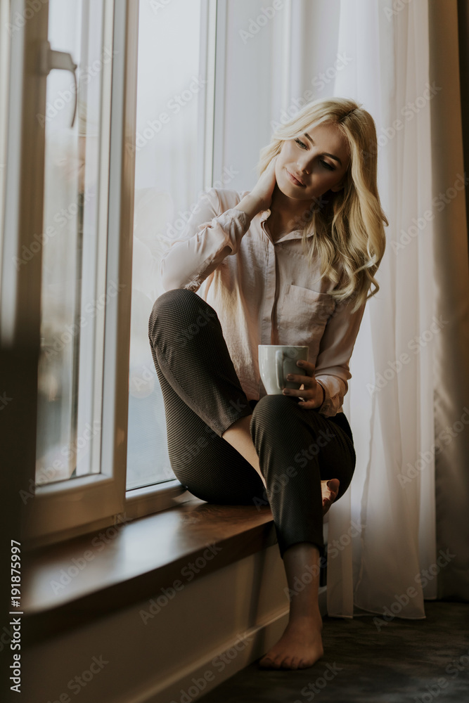 Blonde young woman drinking coffee by window