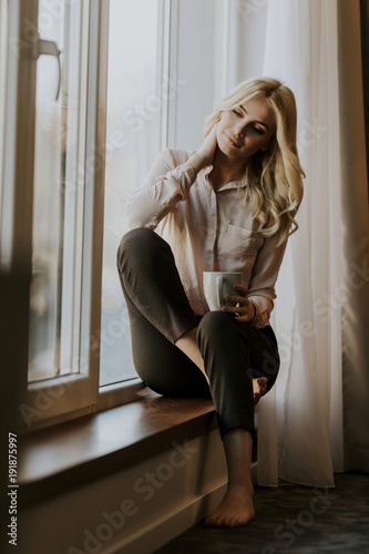 Blonde young woman drinking coffee by window