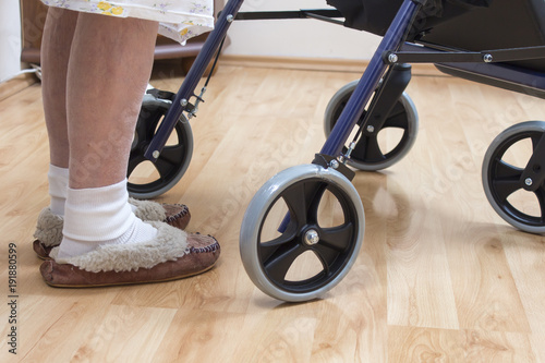 4. Nogi bardzo starej kobiety w białych skarpetkach i pantoflach. Staruszka uczy się chodzić przy pomocy chodzika rehabilitacyjnego.