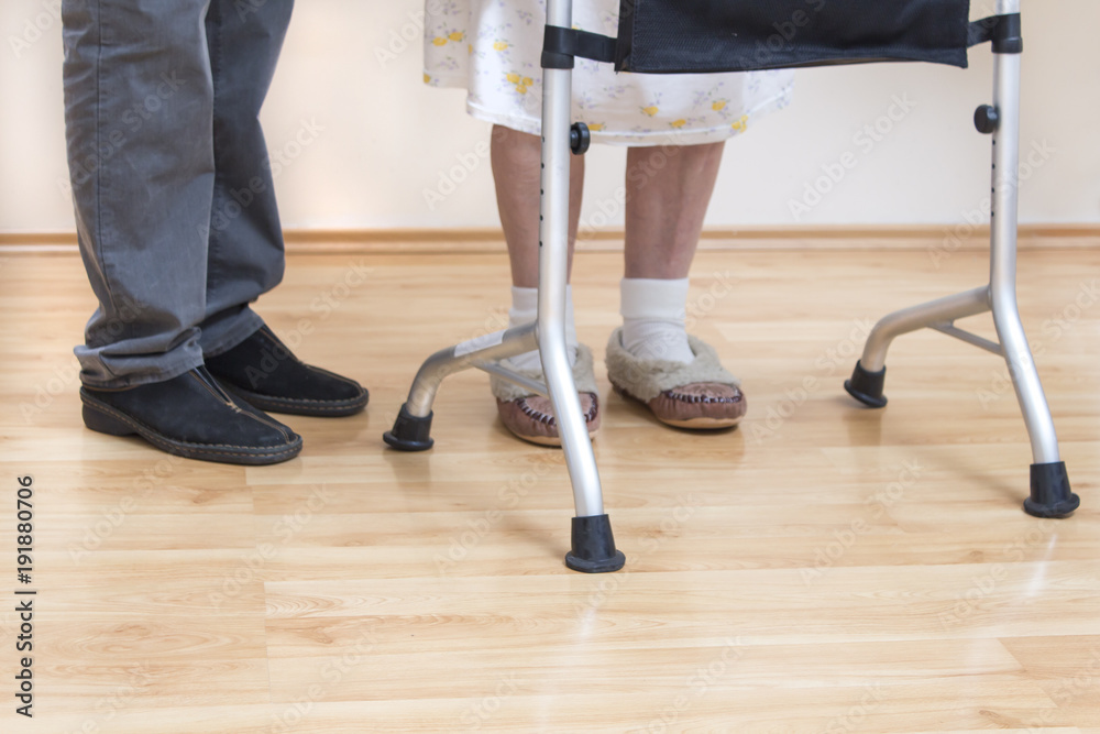4. Nogi bardzo starej kobiety w białych skarpetkach i pantoflach. Staruszka uczy się chodzić przy pomocy chodzika rehabilitacyjnego.