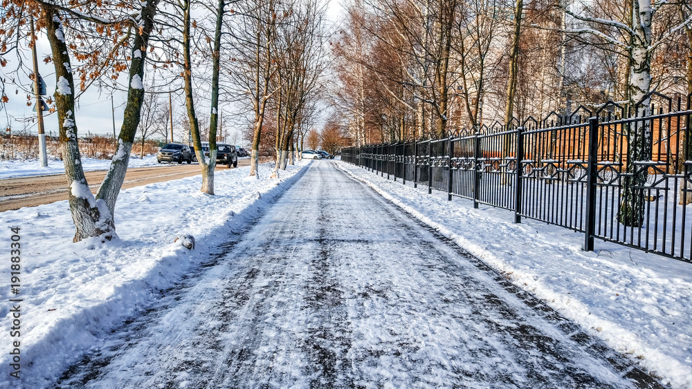 The cleared road in the park, cleaned in winter in the city, the road cleaned by a sunny day. Asphalt in the snow next to the fence. The cleared road in the park, cleaned in winter in the city, the road cleaned by a sunny day. Asphalt in the snow next to the fence.