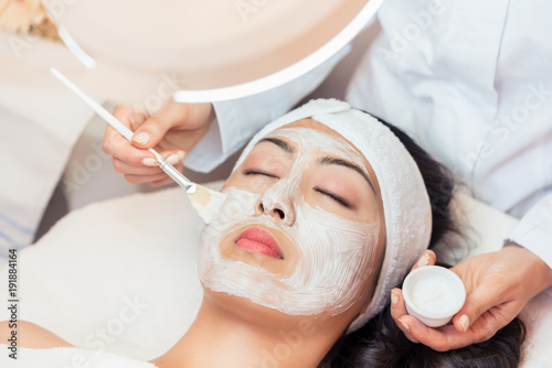 Close-up of the face of a young woman relaxing under the gentle touch of the specialist, applying on her cheeks white facial mask with rejuvenating effects