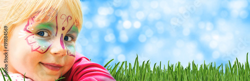 Fototapeta Naklejka Na Ścianę i Meble -  glückliches geschminktes Kind in einem Freizeitpark liegt auf Rasen mit sommerlichem Hintergrund