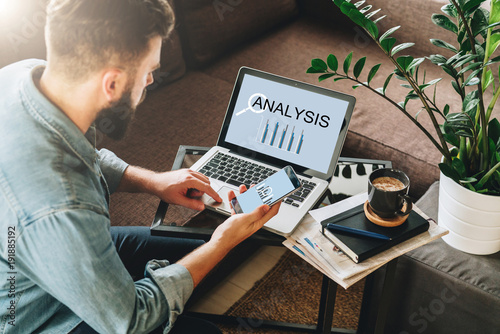 Young man, entrepreneur, freelancer sits at home on couch at coffee table, uses smartphone, working on laptop with inscription analysis on screen.Online marketing,education, e-learning.