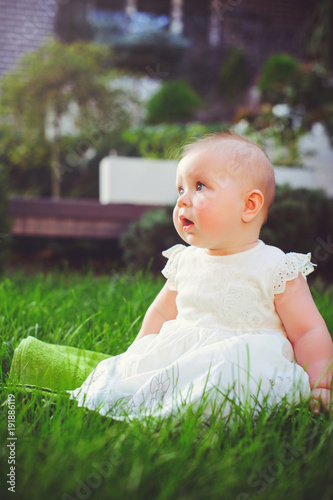 half-year-old child sitting on the grass in the yard, dressed in a white dress rejoices, 6 months. Concept education of children, children's goods