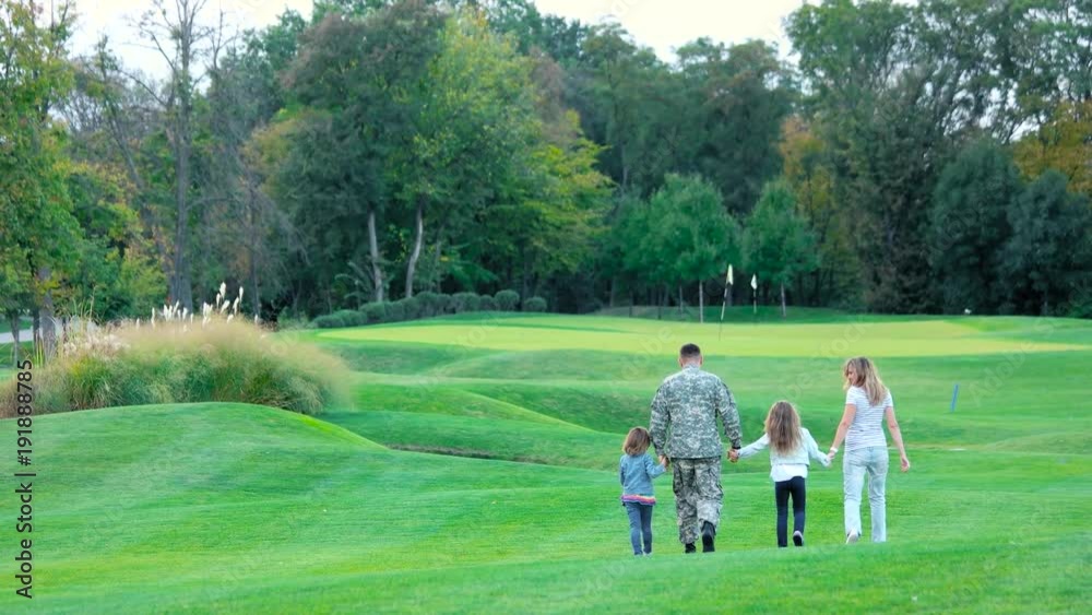 Military man and wife walking and playing with daughters,rear view. Little girls jumping on lawn, back view. Military man and wife walking and playing with daughters,rear view. Little girls jumping on lawn, back view.