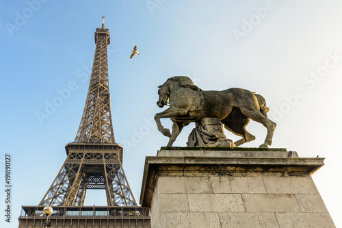 Low angle view of the Eiffel tower in Paris, seen from the Iena bridge against blue sky, with an equestrian stone statue in the foreground.