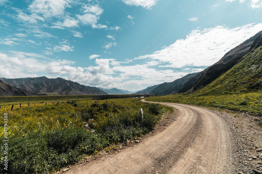 Wide-angle view of unmetalled road twisting among mountains of Altai on sunny summer day surrounded by meadows of native grasses with hills range in a distance, Russia, Kuyus district