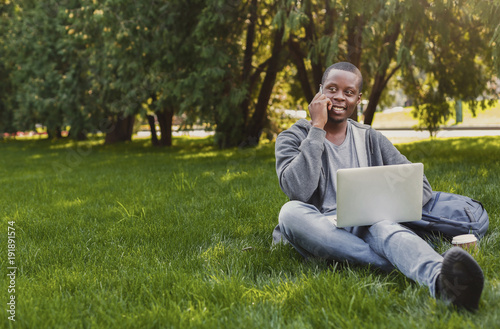 Smiling african-american student talking on phone and using laptop outdoors