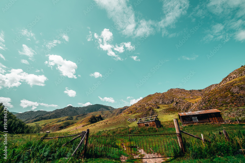 Wide-angle view of summer landscape: old metal gates with feeding ground behind, shepherd's house and abandoned shack, hills range recedes into the distance; Altai mountains, Russia, Kuyus district