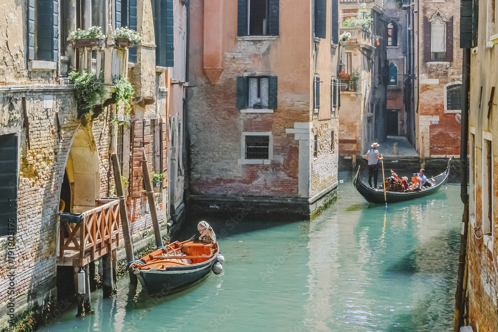 Gondolas on narrow canal and small boat tied next to old red brick house with wooden balcony on narrow canal in Venice, Italy.