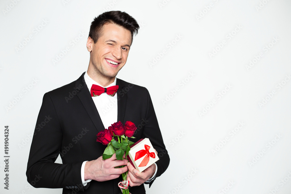 Stylish smiling handsome modern man in a official black suit and a red bow tie is holding a gift box and beautiful red roses in hands. Gift for beloved woman of friend. Stylish smiling handsome modern man in a official black suit and a red bow tie is holding a gift box and beautiful red roses in hands. Gift for beloved woman of friend.