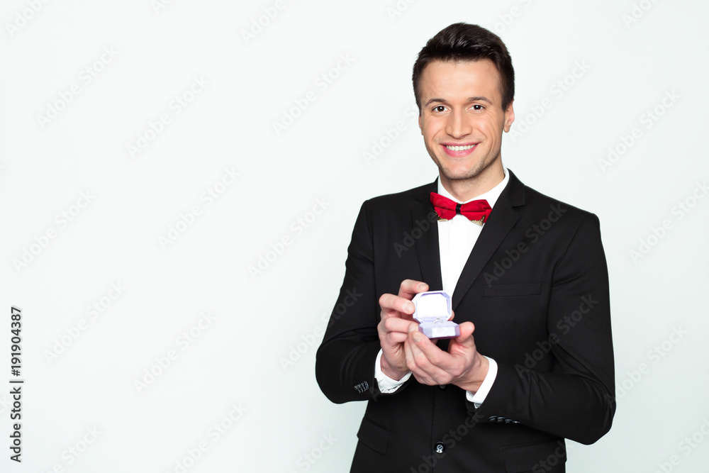 Handsome smiling modern elegant man in a official black suit and a red bow tie is holding a box with an engagement ring and shows in camera. Handsome smiling modern elegant man in a official black suit and a red bow tie is holding a box with an engagement ring and shows in camera.