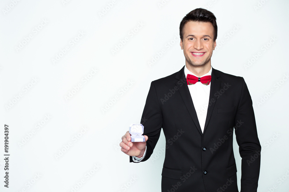 Handsome smiling modern elegant man in a official black suit and a red bow tie is holding a box with an engagement ring and shows in camera. Handsome smiling modern elegant man in a official black suit and a red bow tie is holding a box with an engagement ring and shows in camera.