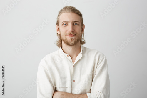 Portrait of positive handsome blond man with beard and moustache, standing with crossed hands in white shirt with slight smile and confident expression. Boyfriend picking flowers for his couple