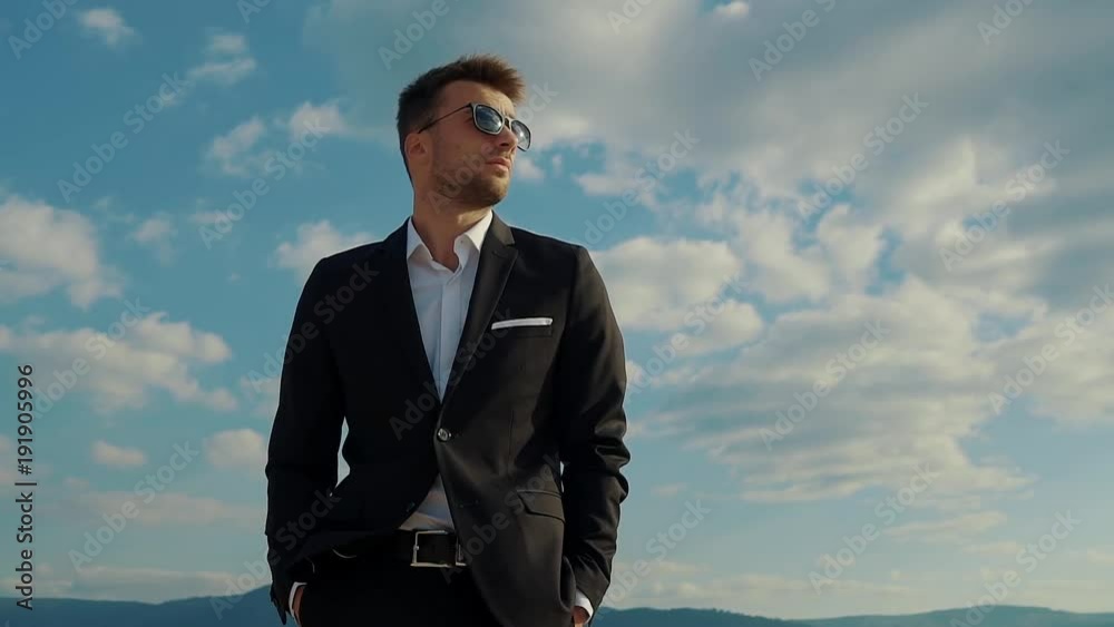 Handsome businessman dressed in a suit standing outdoor in nature. He is looking around in his sunglasses. Handsome businessman dressed in a suit standing outdoor in nature. He is looking around in his sunglasses.