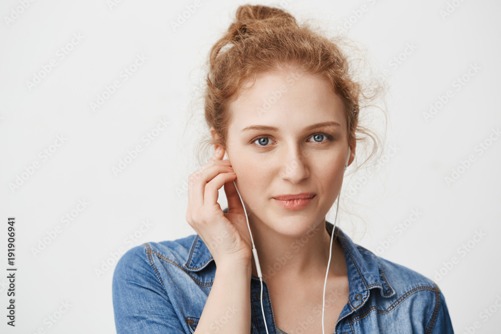 Close-ip portrait of kind gorgeous young redhead girl with blue eyes, wearing earphones while listening music or talking with friend on phone, smiling and standing against gray background. Close-ip portrait of kind gorgeous young redhead girl with blue eyes, wearing earphones while listening music or talking with friend on phone, smiling and standing against gray background.