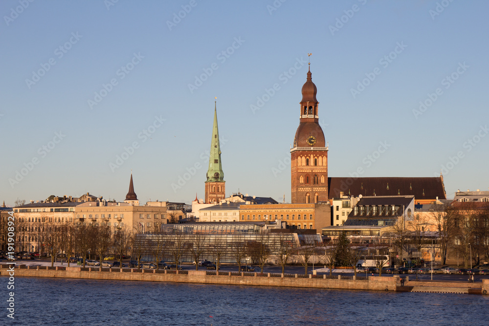 Riga, Latvia. The View Of 11 November Embankment Of River Daugava. The Towers, Steeples Of Riga Cathedral, St. Peter's Church And St. Saviour's Anglican Church In Old Town In Summer Under Blue Sky.