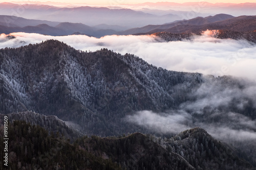 From a viewpoint high in the Great Smokey Mountains National Park stretches a line of mountain ridges to the horizon. It is sunrise and the sky is orange. Low clouds fill the valleys. Misty and foggy