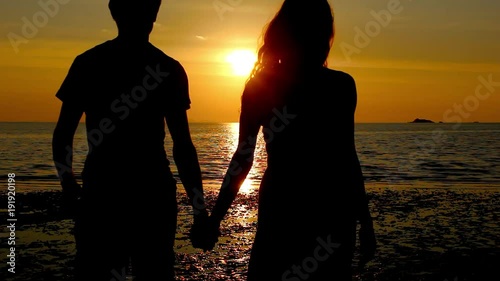 Young couple holding hands walk towards the shore on the beach at sunset in the island of Koh Phangan, Thailand. Valentine Day celebration. Romantic scene, honeymoon love concept