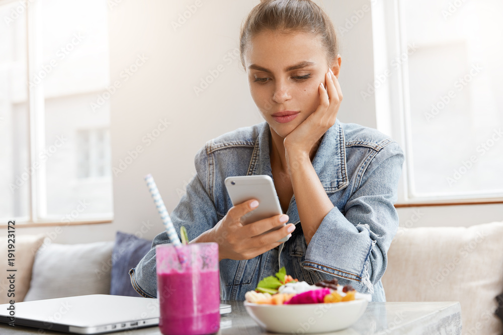 Serious beautiful young woman in denim jacket reads attentively news on webpage via modern smart phone, recreats at cozy restaurant with cocktail and fruit salad, connected to wireless internet. Serious beautiful young woman in denim jacket reads attentively news on webpage via modern smart phone, recreats at cozy restaurant with cocktail and fruit salad, connected to wireless internet.