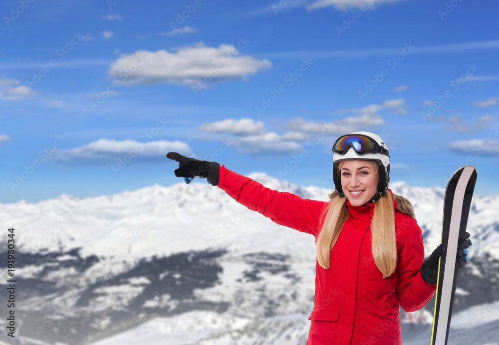 Skier on the background of snow-capped mountains. Attractive blonde in a red ski suit with snowboard in hands shows on snowy mountains.