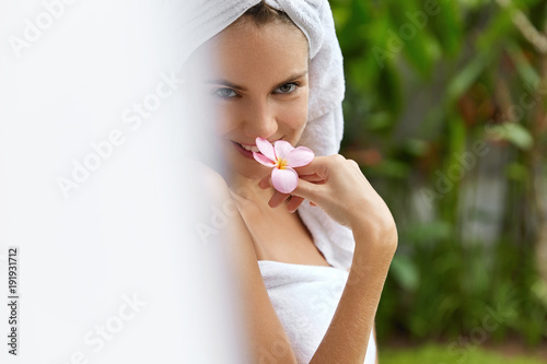 Pretty young female wrapped in towels  has pure healthy soft skin  smells odour orchid  being pleased after aromatherapy procedure  looks happily and shy into camera. Beauty and care concept