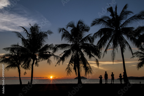 A group of people witnessing the sun setting over the horizon by the esplanade on a clear sky day.