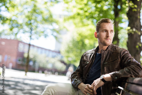 Lonely man sitting down on a bench outdoors on a sunny day, wearing a blue shirt and brown leather jacket. Looking away from camera.
