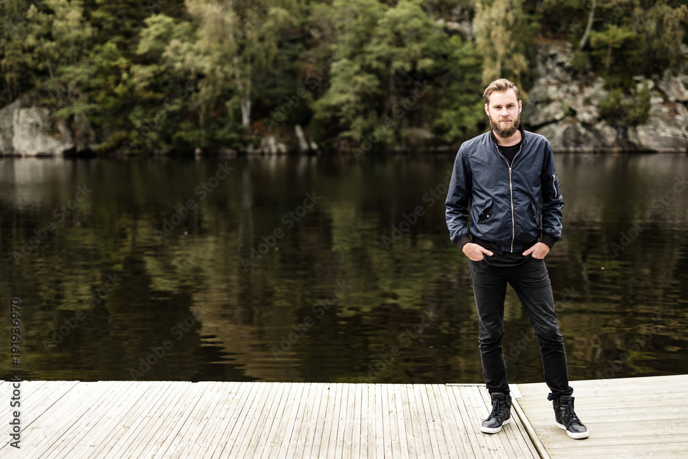 Fototapeta premium One bearded man wearing a blue jacket, standing in front of a lake and trees on a moody day.