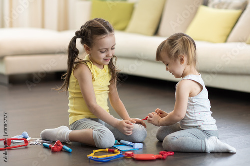 Little child girl playing doctor with her younger toddler sister in living room