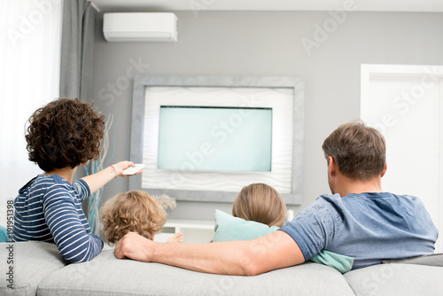Back view portrait of happy family with two children watching TV sitting on sofa in living room and turning on modern television set with remote controller.