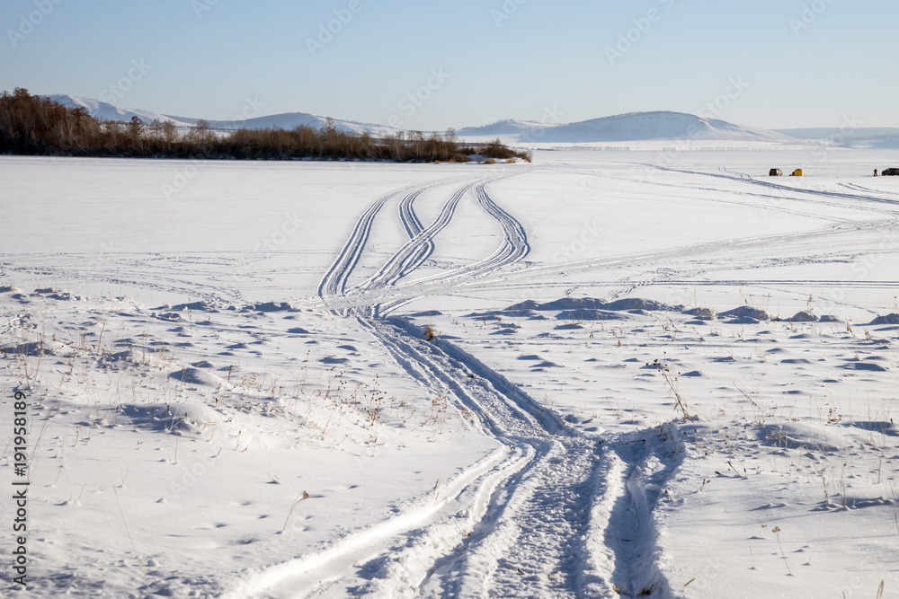Trace from snowmobile on a snowy lake with fishermen in the background 