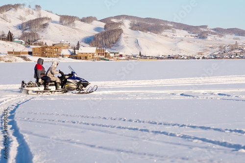 The snowmobile with unidentified fishermen is riding along the snowy lake against the backdrop of the village, in the winter