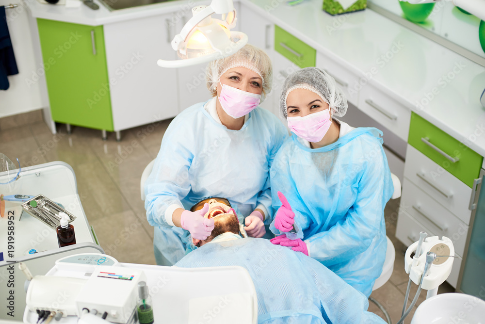 Photo from above doctor woman and her assistant dressed in a one-time uniform work with a male patient in a modern dentistry office and shows gesture class Photo from above doctor woman and her assistant dressed in a one-time uniform work with a male patient in a modern dentistry office and shows gesture class