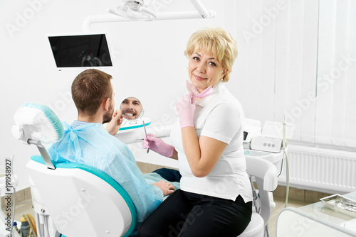 Beautiful female dentist is looking to the camera while holding a mirror for a male client of a dentistry who is smiling checking his teeth.