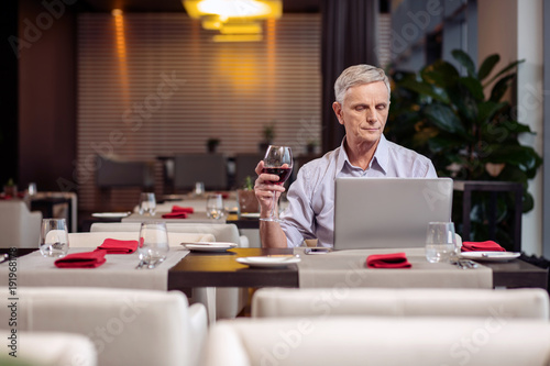 Pleasant evening. Concentrated earnest mature man holding wine while staring at the laptop and resting at the restaurant