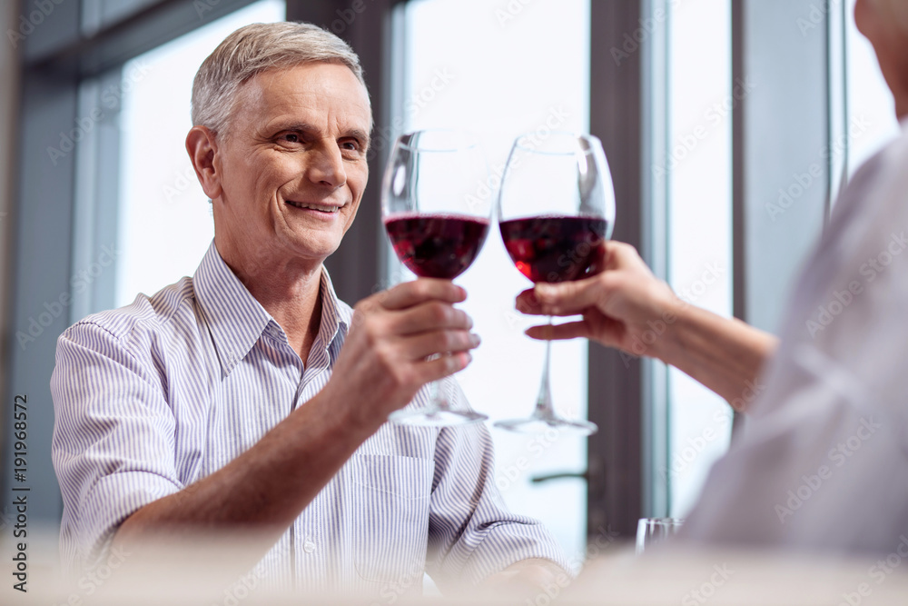 Be healthy. Happy merry mature man rising glass of wine while gazing straight and grinning
