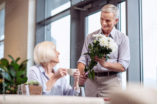 Beautiful as you. Joyful jolly mature man staying with flowers while smiling and woman drinking coffee
