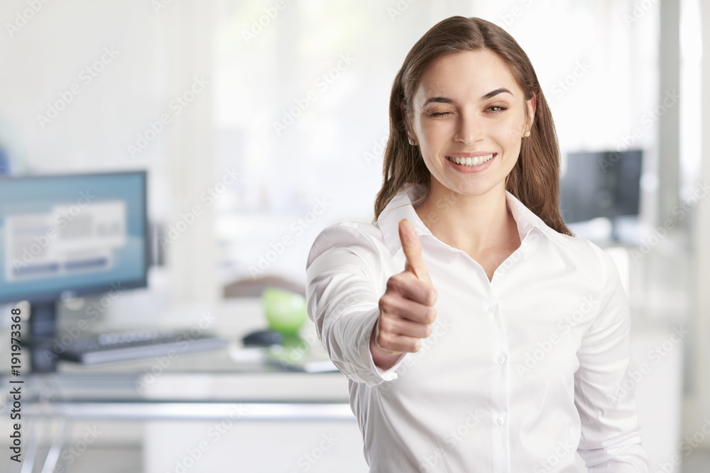 Beautiful young businesswoman with toothy smile giving thumbs up while standing at the office. 