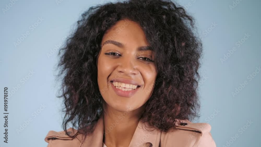 Young african american woman posing over blue background. Trendy and urban style. Afro curly hair, snow-white smile, healthy lifestyle. Slow motion. Young african american woman posing over blue background. Trendy and urban style. Afro curly hair, snow-white smile, healthy lifestyle. Slow motion.