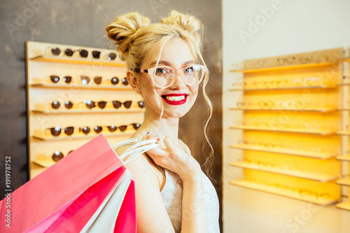 Close up emotional portrait of caucasian blond female shopaholic in eyeglasses with cute hairstyle holding paper bag in front of display at eye wear shop. Consumerism, sale and pleasure concept. photo