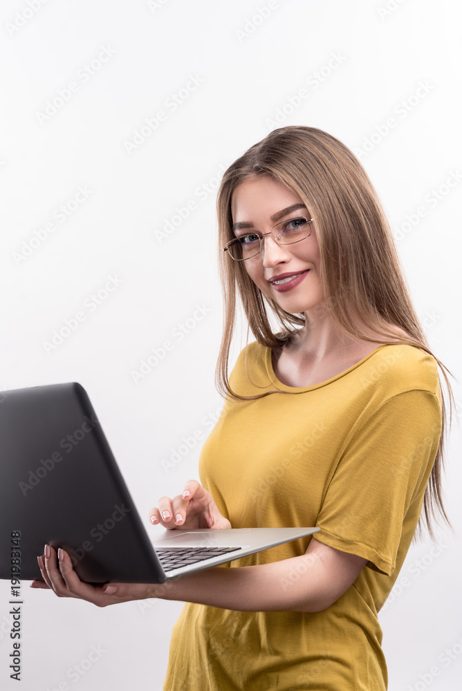 Implementing ideas. Charming young woman in eyeglasses holding a laptop and working on it while smiling at camera and posing isolated on a white background Implementing ideas. Charming young woman in eyeglasses holding a laptop and working on it while smiling at camera and posing isolated on a white background