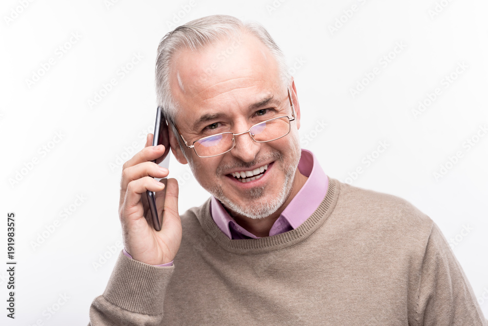 Nice chat. The close up of a handsome grey-haired senior man in eyeglasses talking on the phone and smiling at the camera while posing isolated on a white background Nice chat. The close up of a handsome grey-haired senior man in eyeglasses talking on the phone and smiling at the camera while posing isolated on a white background